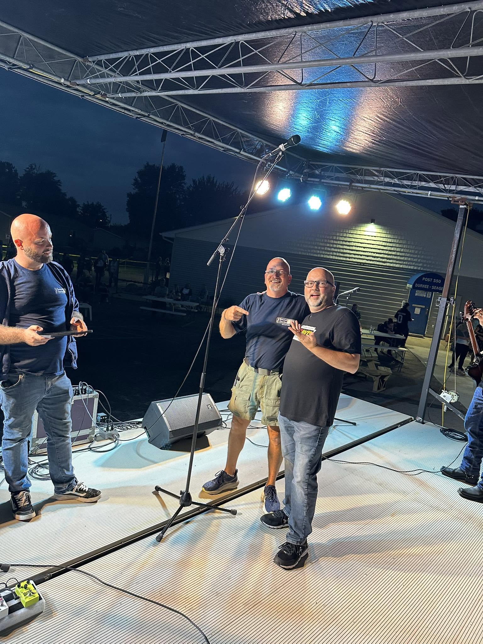 Three bald men on stage at night, one making hang loose gesture